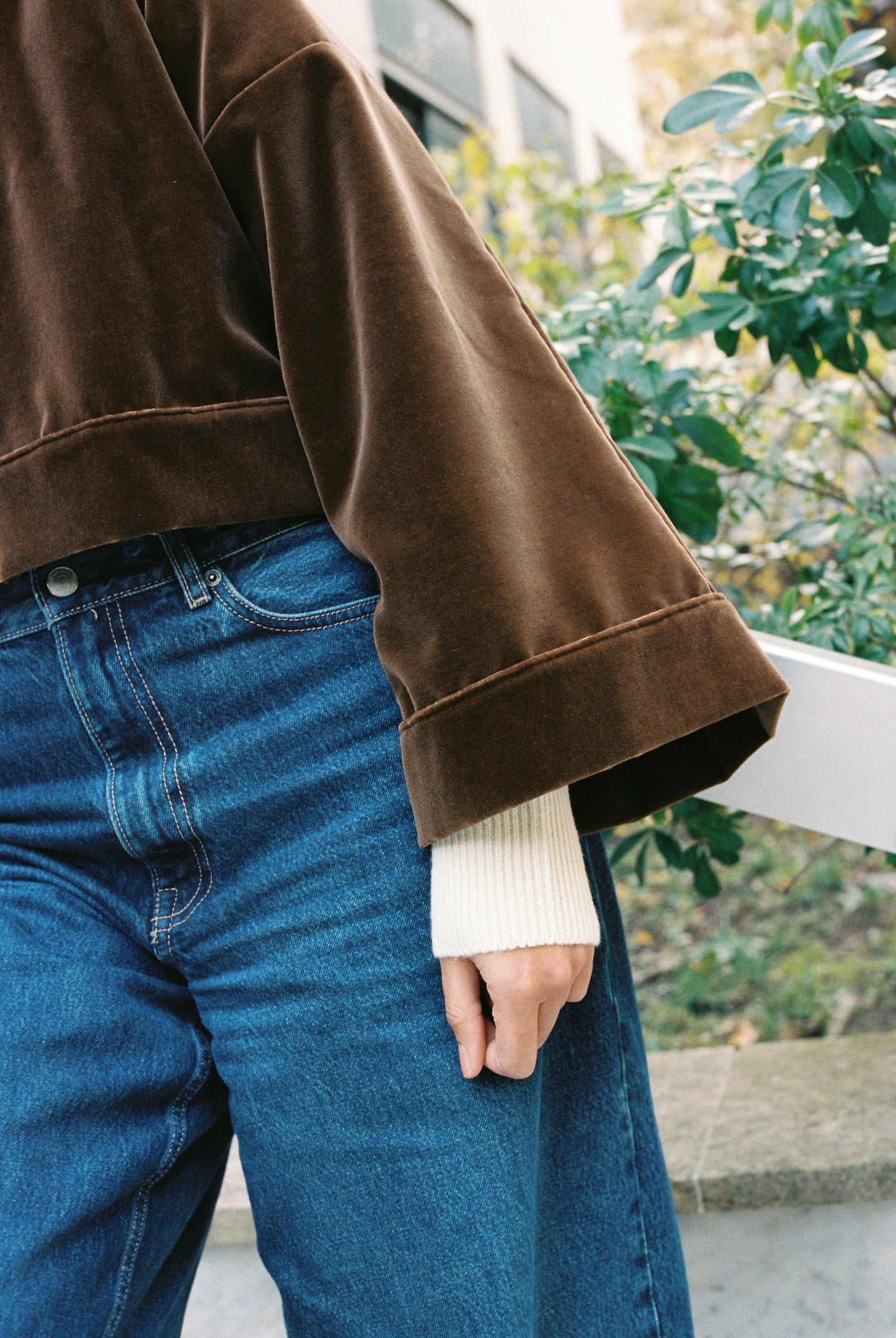 Femme portant le kimono court BRADY en velours de coton marron café Atelier Sarita avec poches intégrées. Kimono fabriqué à la main à Paris dans un velours 100 % coton issu de deadstocks. Pièce chaude et texturée, au tombé souple, photographiée en extérieur avec un look hivernal composé d’un pull écru et d’un jean, mettant en valeur la douceur et la profondeur du velours marron.