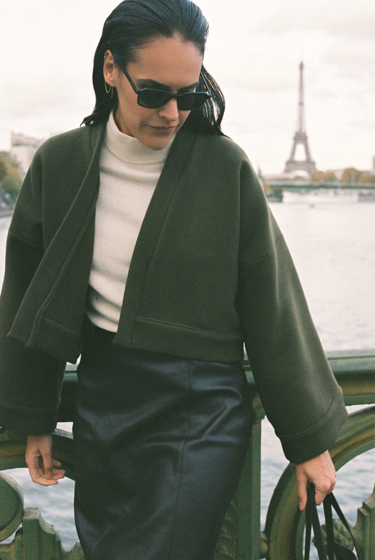 Femme portant le kimono court JOUFFROY Atelier Sarita en laine et cachemire kaki, coupe crop, avec finitions minimalistes et poches intérieures. La silhouette est photographiée sur un pont parisien avec vue sur la Tour Eiffel, mettant en valeur un kimono artisanal confectionné à la main à Paris, pensé pour l’hiver.