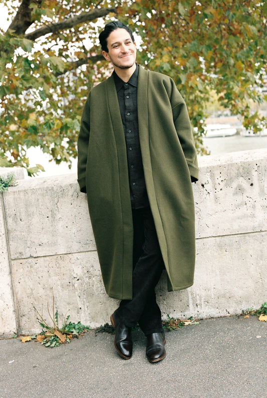 Homme portant le kimono long VIVIENNE en laine et cachemire kaki d’Atelier Sarita. Kimono oversize et structuré, fabriqué à la main à Paris dans un lainage italien issu de stocks dormants de maison de luxe. Pièce enveloppante et élégante, photographiée en extérieur avec la tour Eiffel en arrière-plan, mettant en valeur la douceur, la tenue et la chaleur du tissu.
