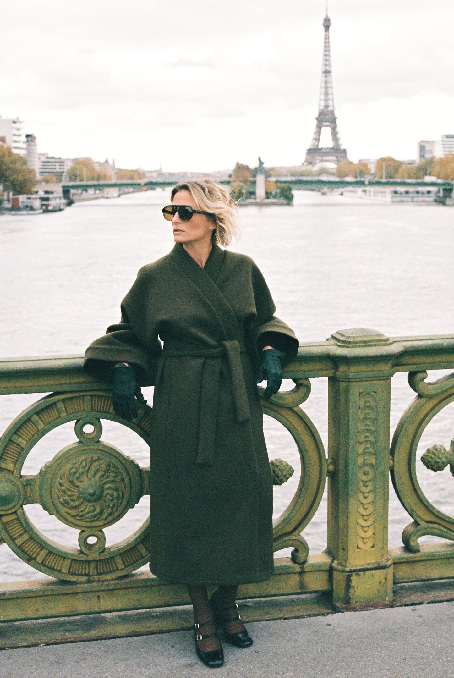 Femme portant le kimono long VIVIENNE en laine et cachemire kaki d’Atelier Sarita. Kimono oversize et structuré, fabriqué à la main à Paris dans un lainage italien issu de stocks dormants de maison de luxe. Pièce enveloppante et élégante, photographiée en extérieur avec la tour Eiffel en arrière-plan, mettant en valeur la douceur, la tenue et la chaleur du tissu.