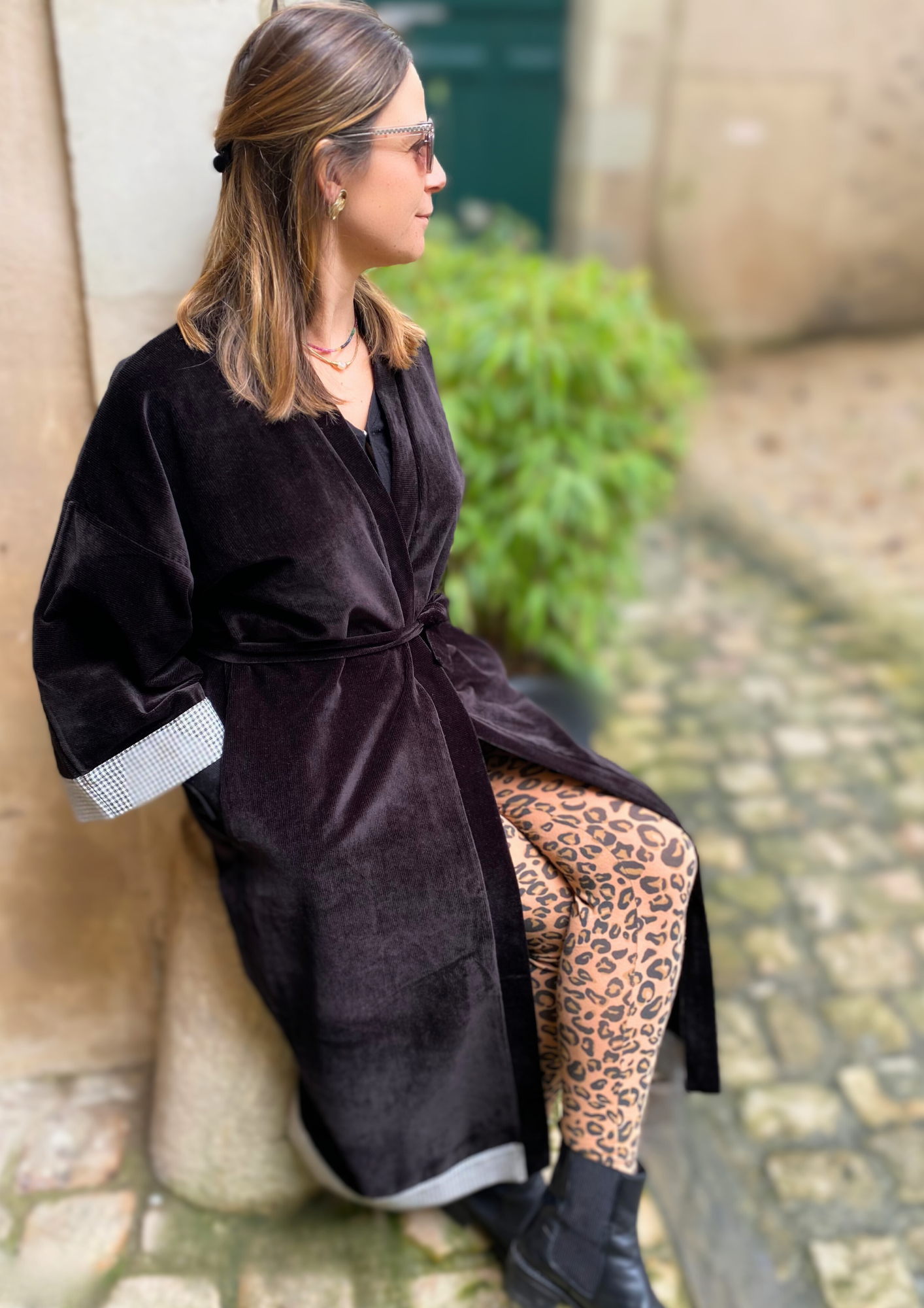 Jeune femme assise en dans une cour vêtue d'un Kimono long en velours noir avec détails laine vichy aux poignets et à l'ourlet, coupe fluide et manches larges, ceinturé.