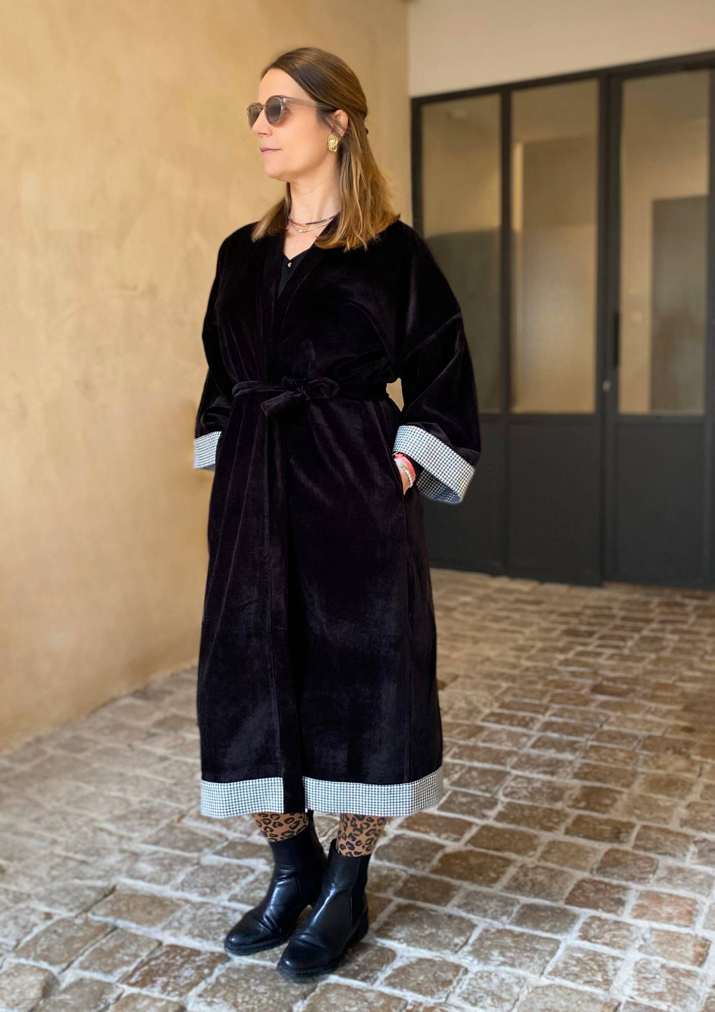 Jeune femme dans une cour vêtue d'un Kimono long en velours noir avec détails laine vichy aux poignets et à l'ourlet, coupe fluide et manches larges, ceinturé.