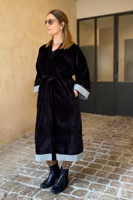Jeune femme dans une cour vêtue d'un Kimono long en velours noir avec détails laine vichy aux poignets et à l'ourlet, coupe fluide et manches larges, ceinturé.