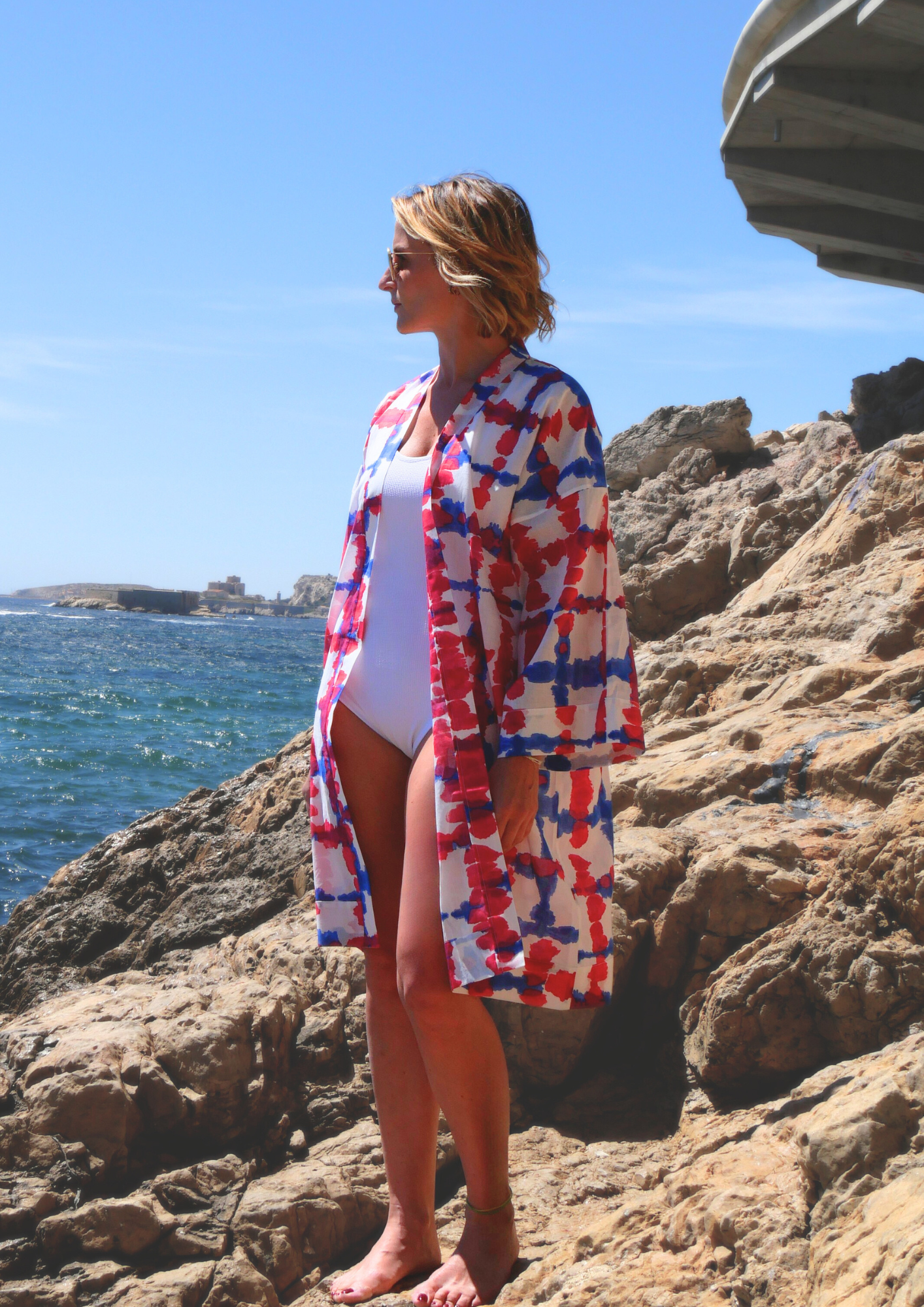 Femme portant un kimono mi-long bleu et fuchsia Atelier Sarita, en voile de coton léger, sur un rocher face à la mer.