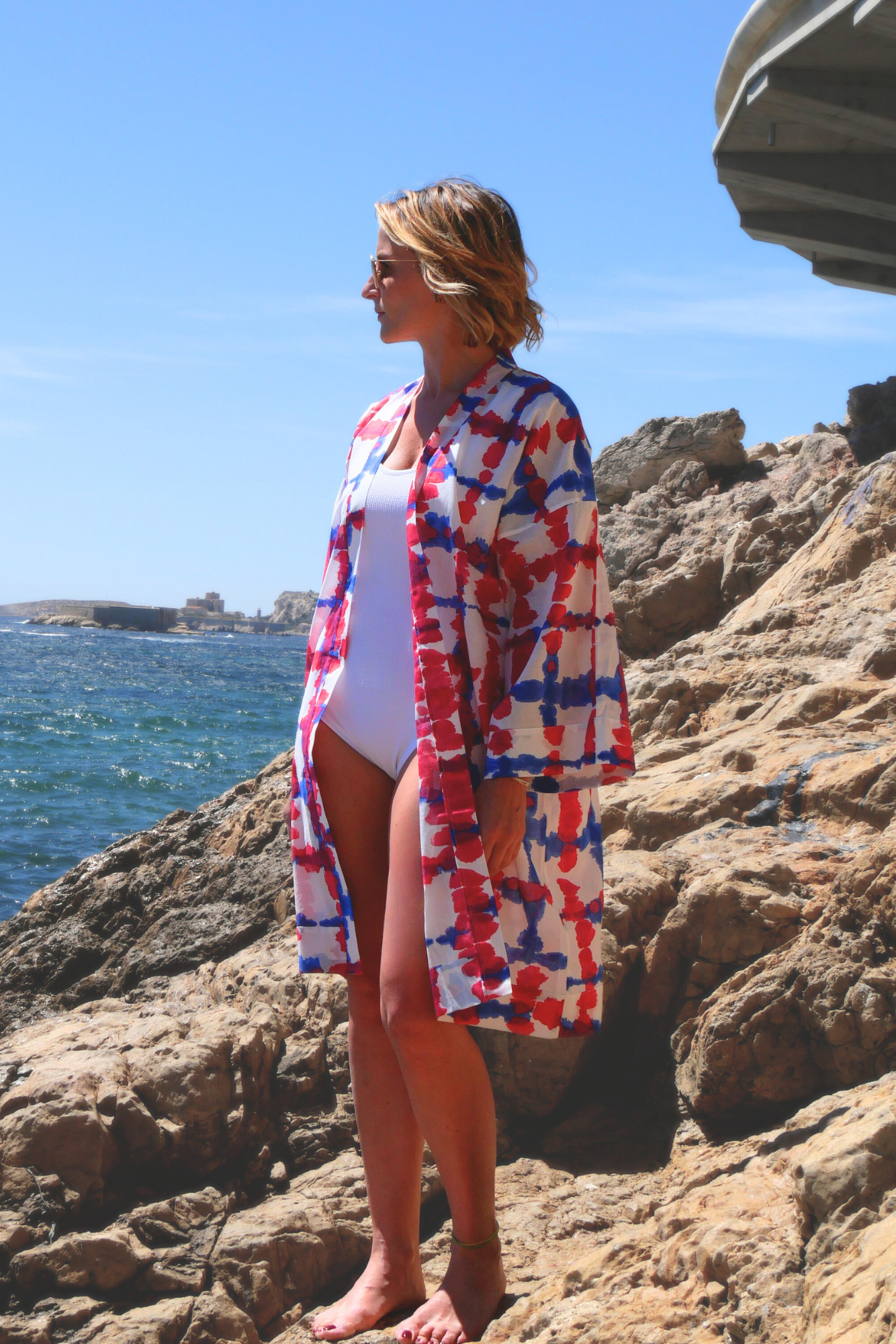 Femme portant un kimono mi-long bleu et fuchsia Atelier Sarita, en voile de coton léger, sur un rocher face à la mer.
