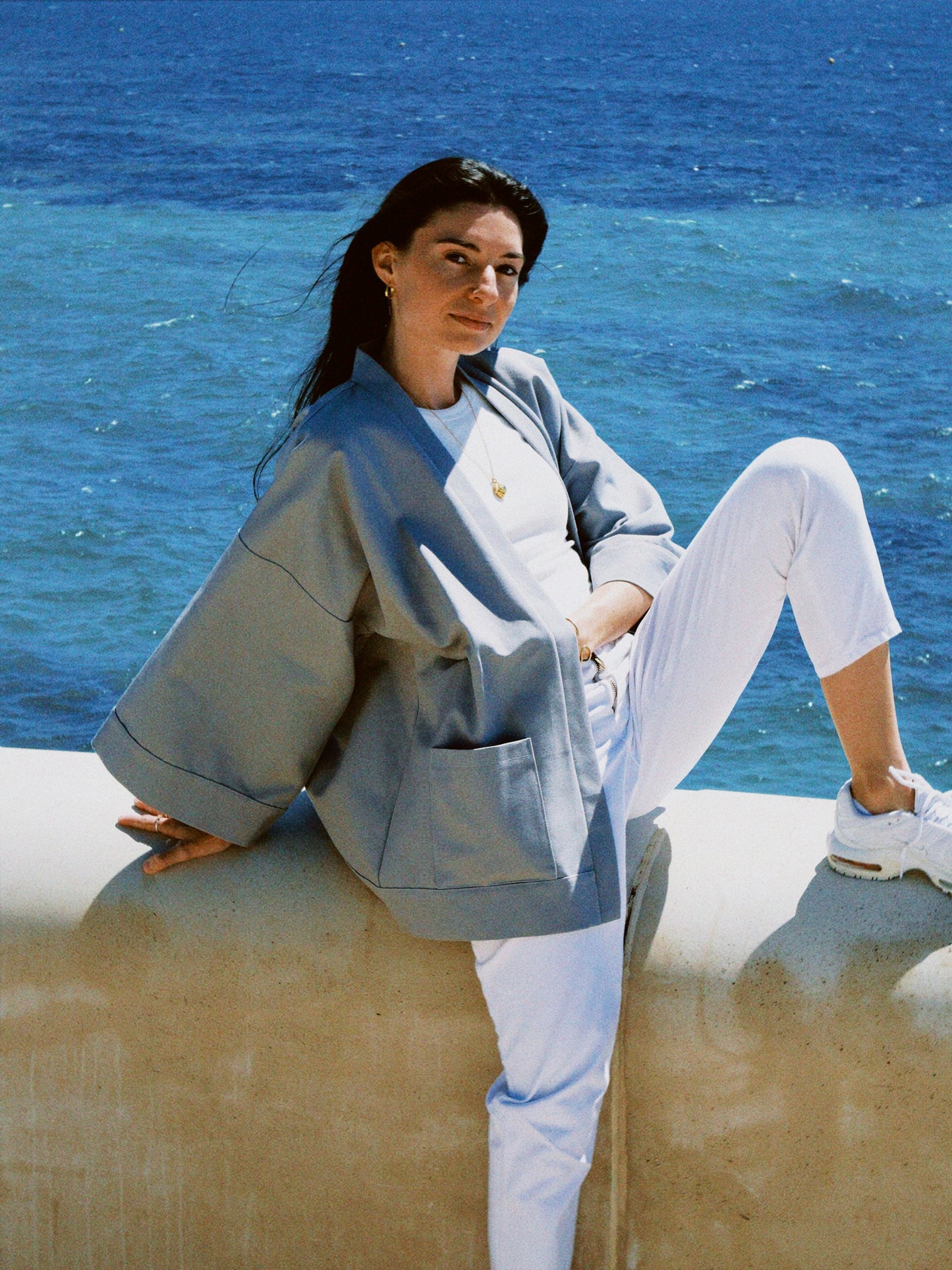 Femme portant un kimono gris en coton biologique, coupe ample avec ceinture, photographiée en bord de mer. Pièce confectionnée à la main à Paris, par Atelier Sarita.