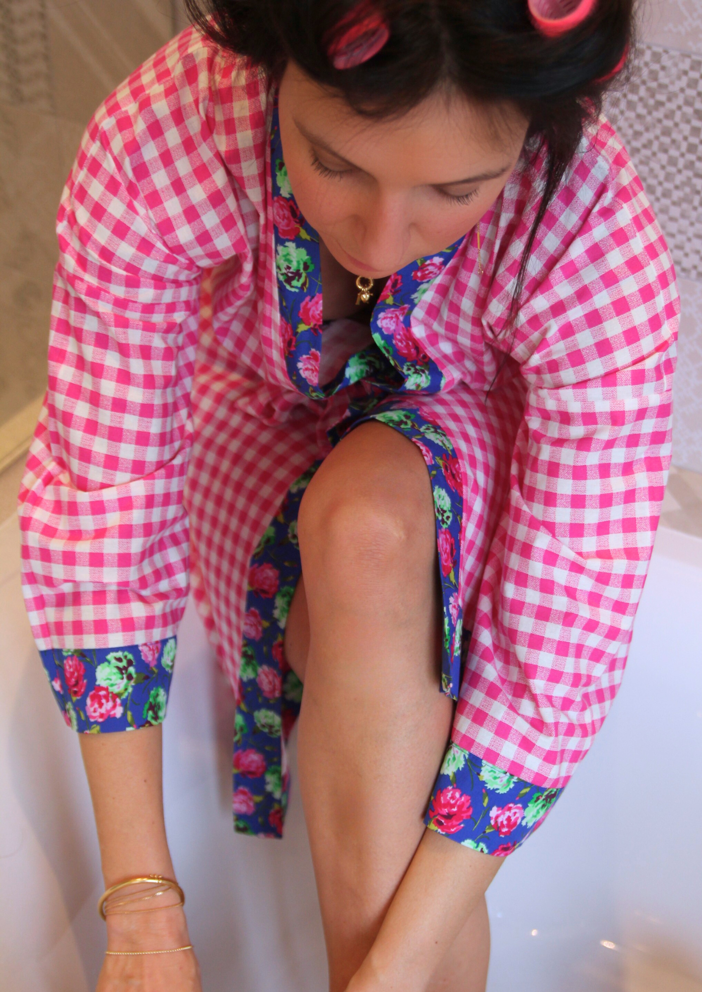 Femme dans salle de bain vêtue d'un kimono mi-long en tissu vichy rose avec col et finitions fleuries, manches larges et coupe fluide.