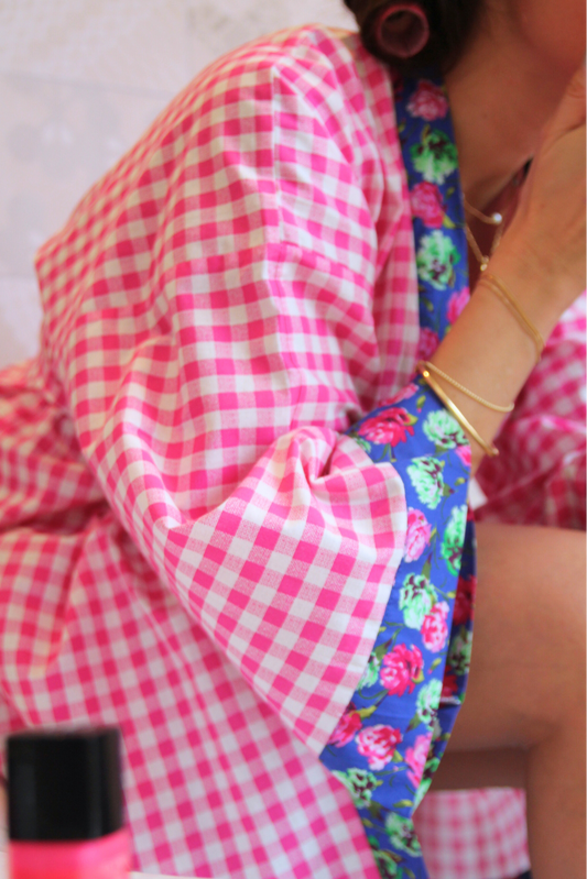 Femme dans salle de bain vêtue d'un kimono mi-long en tissu vichy rose avec col et finitions fleuries, manches larges et coupe fluide.