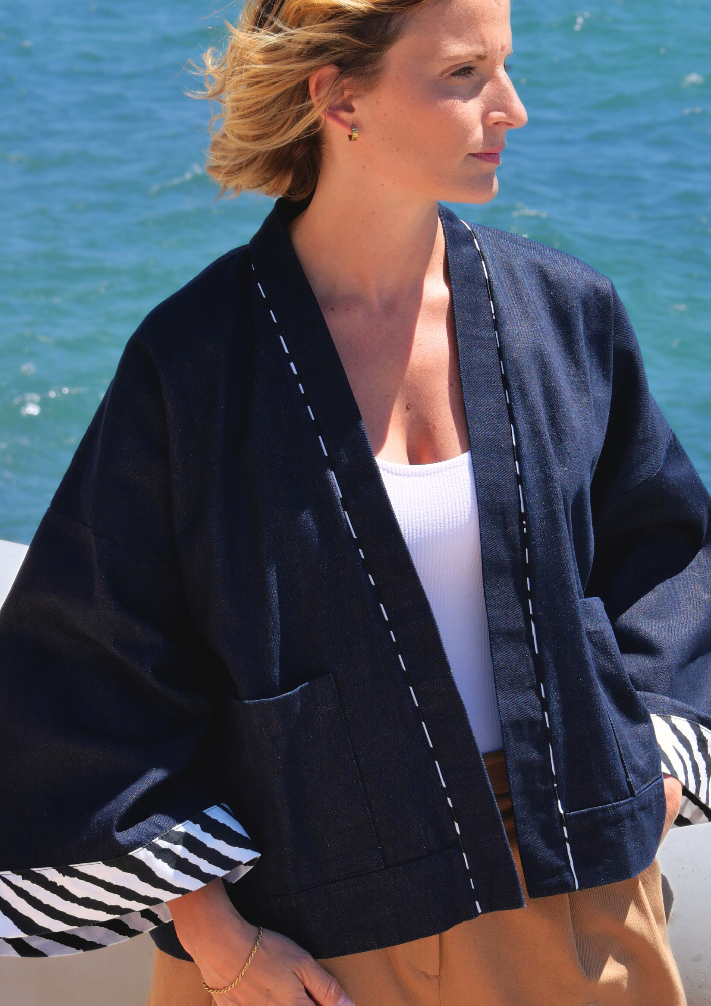 Femme portant un kimono court en denim bleu avec finitions zébrées à l'encolure et aux manches, modèle confectionné à la main par Atelier Sarita, photographiée face à la mer.