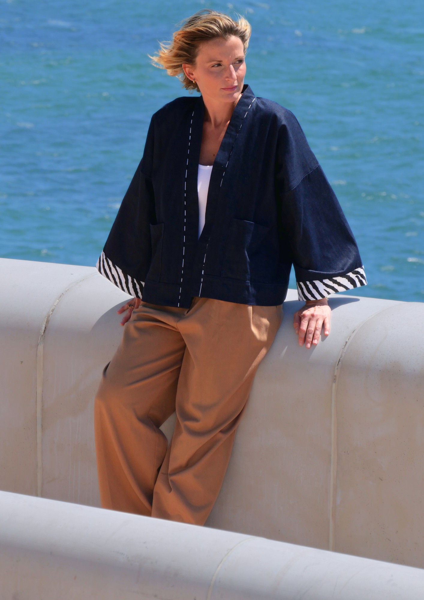 Femme portant un kimono court en denim bleu avec finitions zébrées à l'encolure et aux manches, modèle confectionné à la main par Atelier Sarita, photographiée face à la mer.