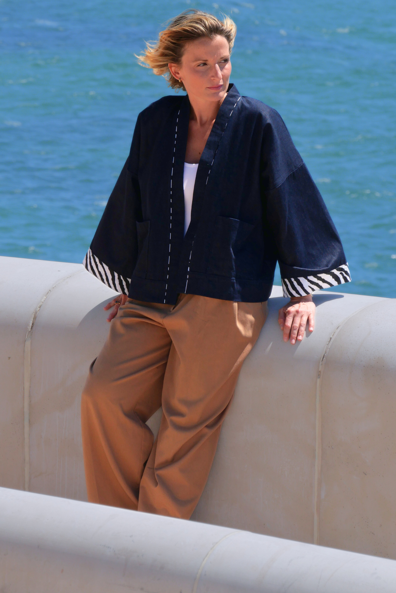 Femme portant un kimono court en denim bleu avec finitions zébrées à l'encolure et aux manches, modèle confectionné à la main par Atelier Sarita, photographiée face à la mer.