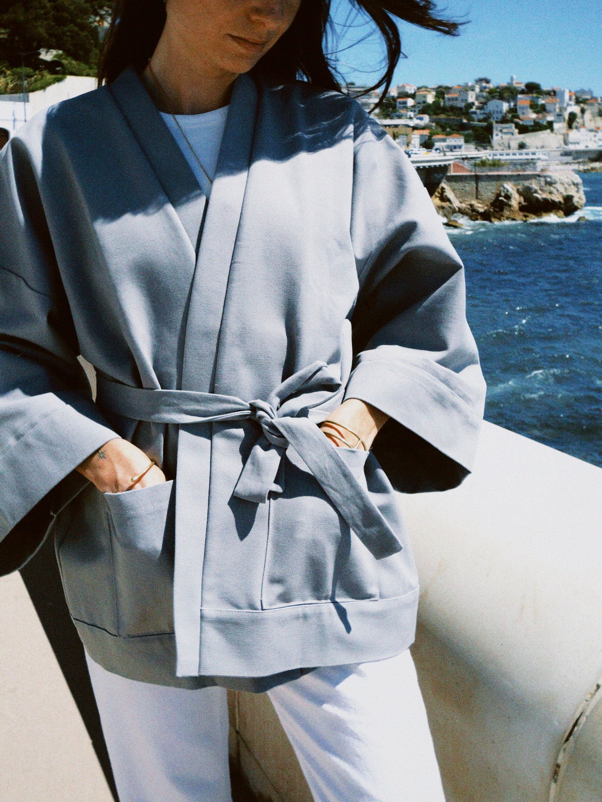 Femme portant un kimono gris en coton biologique, coupe ample avec ceinture, photographiée en bord de mer. Pièce confectionnée à la main à Paris, par Atelier Sarita.