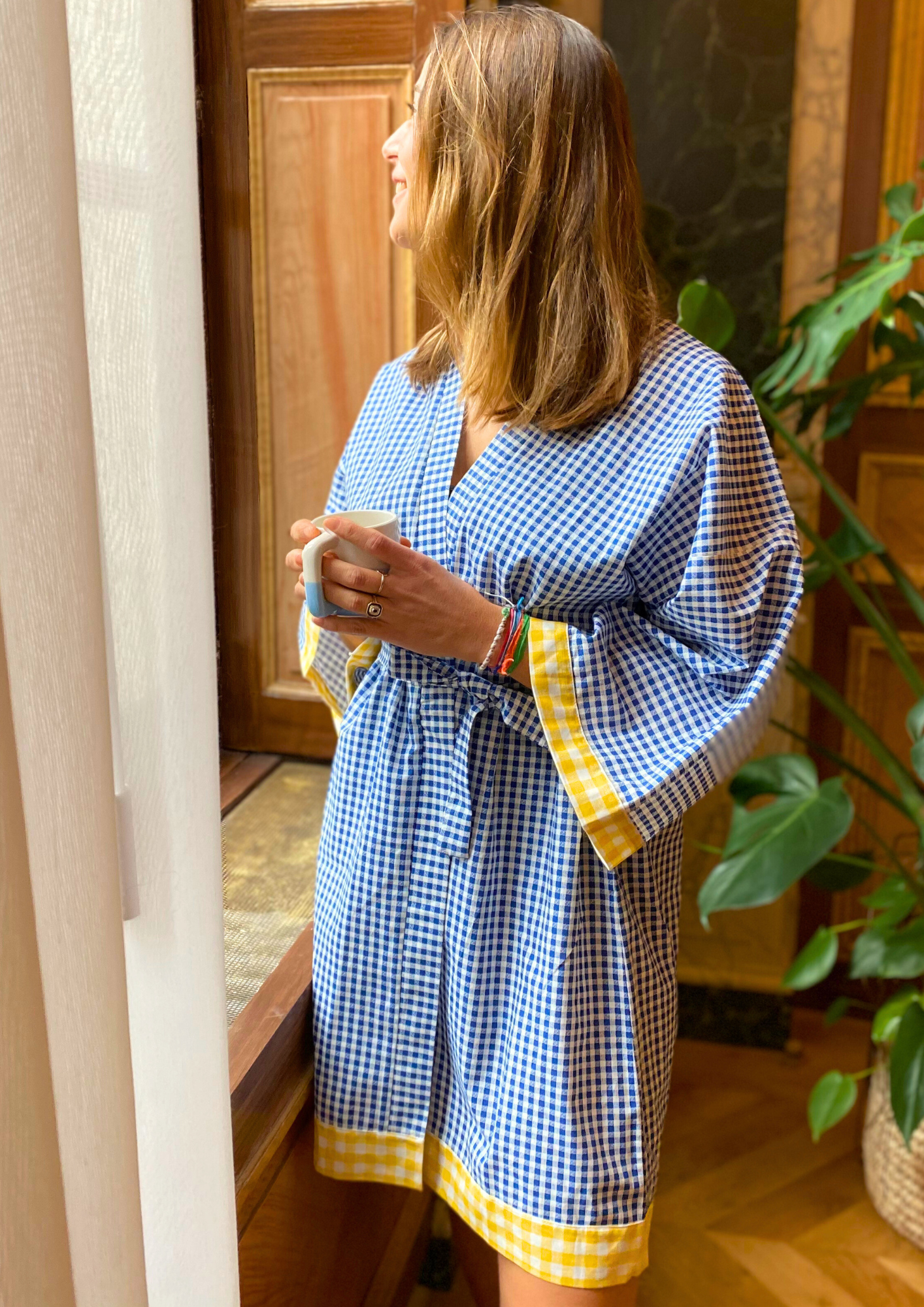 Jeune femme en kimono mi-long en coton vichy bleu avec finitions jaunes, ceinture ajustable, manches larges et coupe fluide, regarde par la fenêtre en busant son café.