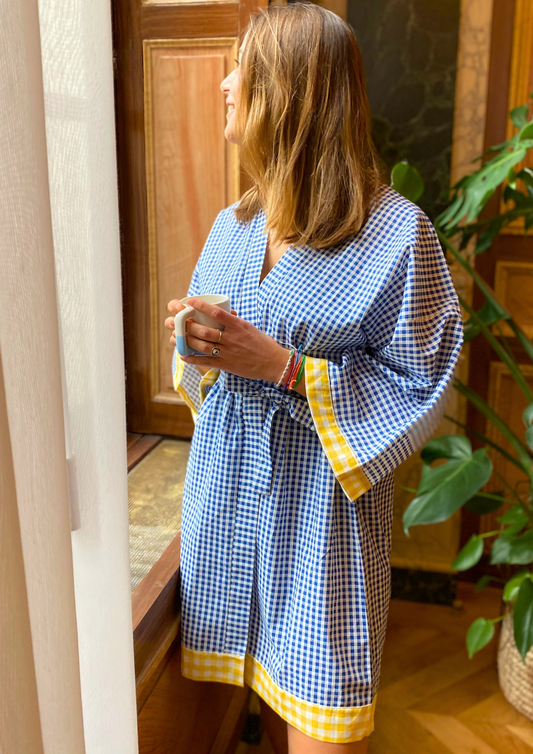 Jeune femme en kimono mi-long en coton vichy bleu avec finitions jaunes, ceinture ajustable, manches larges et coupe fluide, regarde par la fenêtre en busant son café.