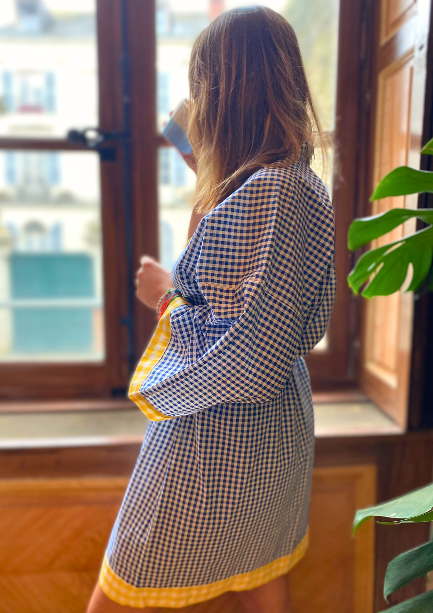 Jeune femme en kimono mi-long en coton vichy bleu avec finitions jaunes, ceinture ajustable, manches larges et coupe fluide, regarde par la fenêtre en busant son café.