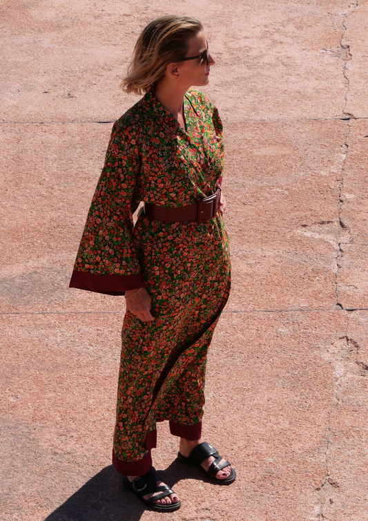 Femme portant un kimono long fleuri en coton avec finitions bordeaux, ceinturé à la taille, debout sur une terrasse ensoleillée – création Atelier Sarita, confectionnée à la main à Paris.