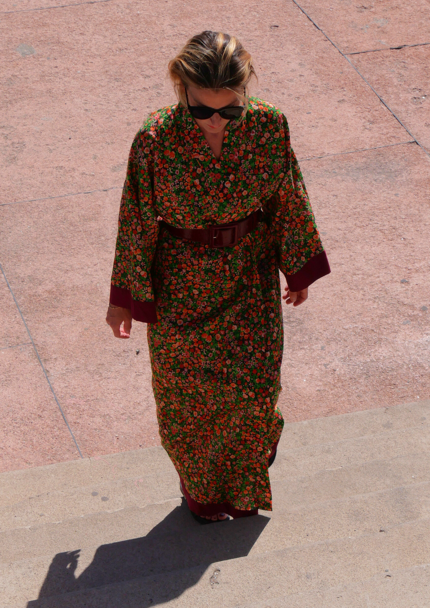 Femme portant un kimono long fleuri en coton avec finitions bordeaux, ceinturé à la taille, debout sur une terrasse ensoleillée – création Atelier Sarita, confectionnée à la main à Paris.