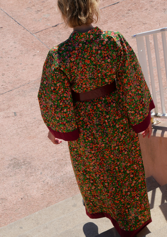 Femme portant un kimono long fleuri en coton avec finitions bordeaux, ceinturé à la taille, debout sur une terrasse ensoleillée – création Atelier Sarita, confectionnée à la main à Paris.