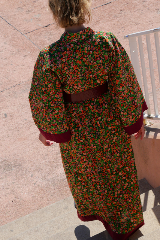 Femme portant un kimono long fleuri en coton avec finitions bordeaux, ceinturé à la taille, debout sur une terrasse ensoleillée – création Atelier Sarita, confectionnée à la main à Paris.