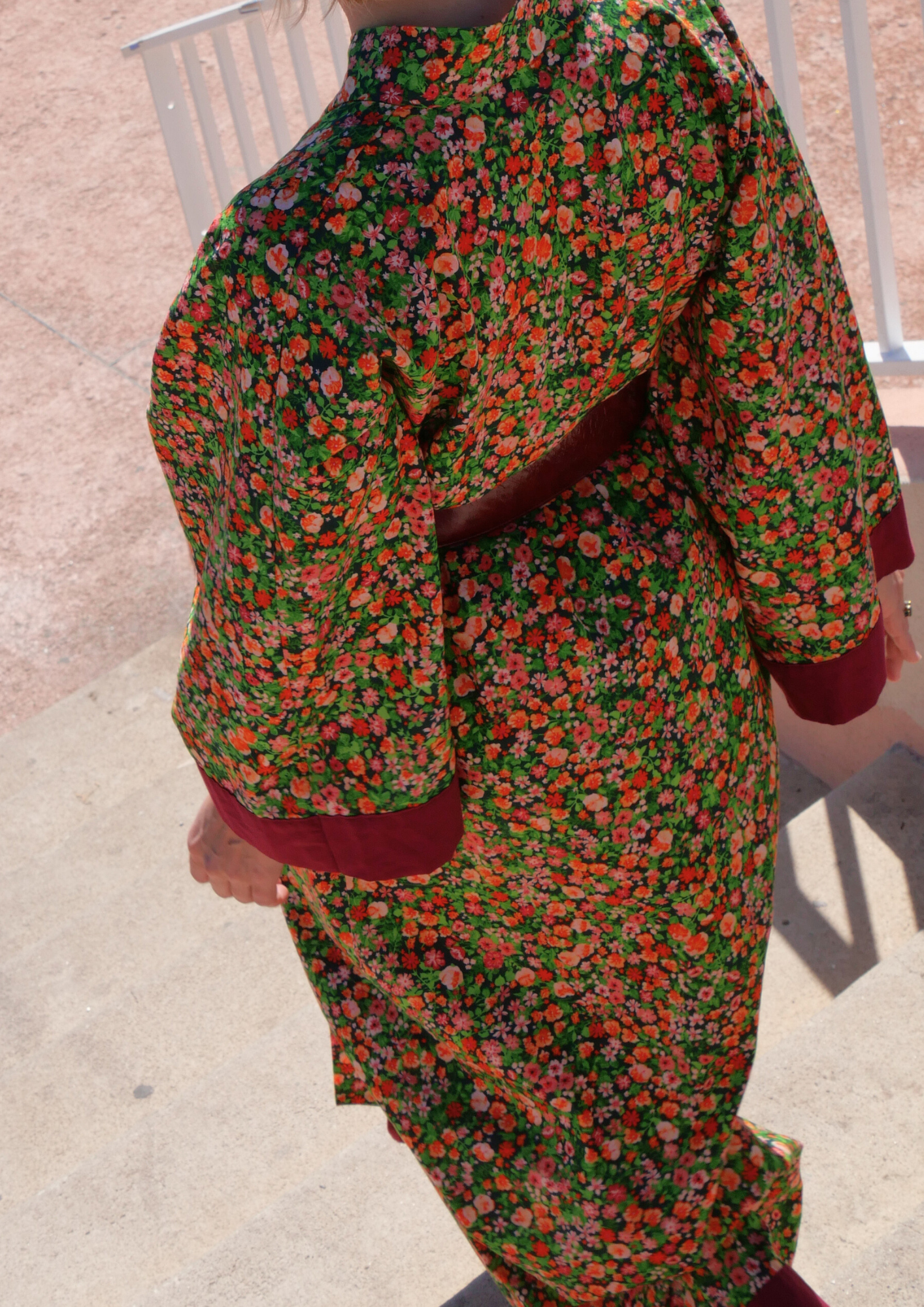 Femme portant un kimono long fleuri en coton avec finitions bordeaux, ceinturé à la taille, debout sur une terrasse ensoleillée – création Atelier Sarita, confectionnée à la main à Paris.
