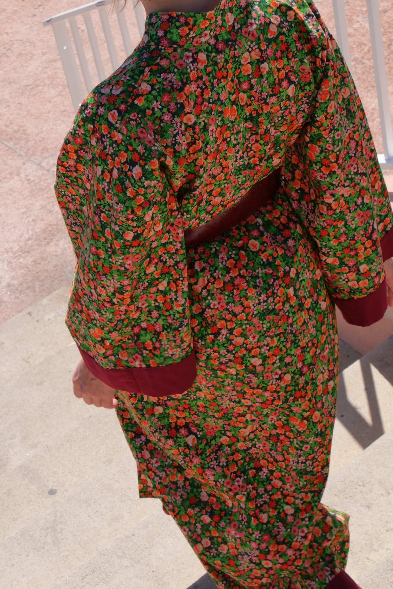 Femme portant un kimono long fleuri en coton avec finitions bordeaux, ceinturé à la taille, debout sur une terrasse ensoleillée – création Atelier Sarita, confectionnée à la main à Paris.