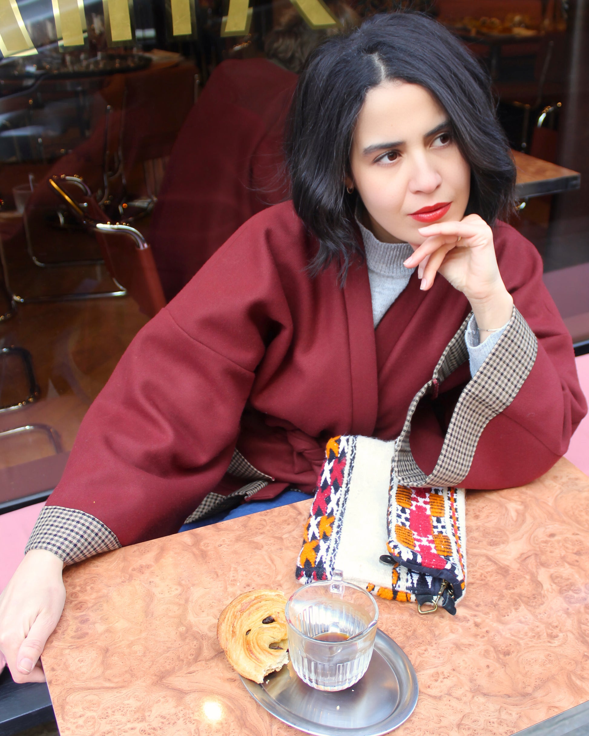 Jeune femme à la terrasse d'un café parisien, portant un kimono en laine bordeaux et finitions en laine pied de poule.