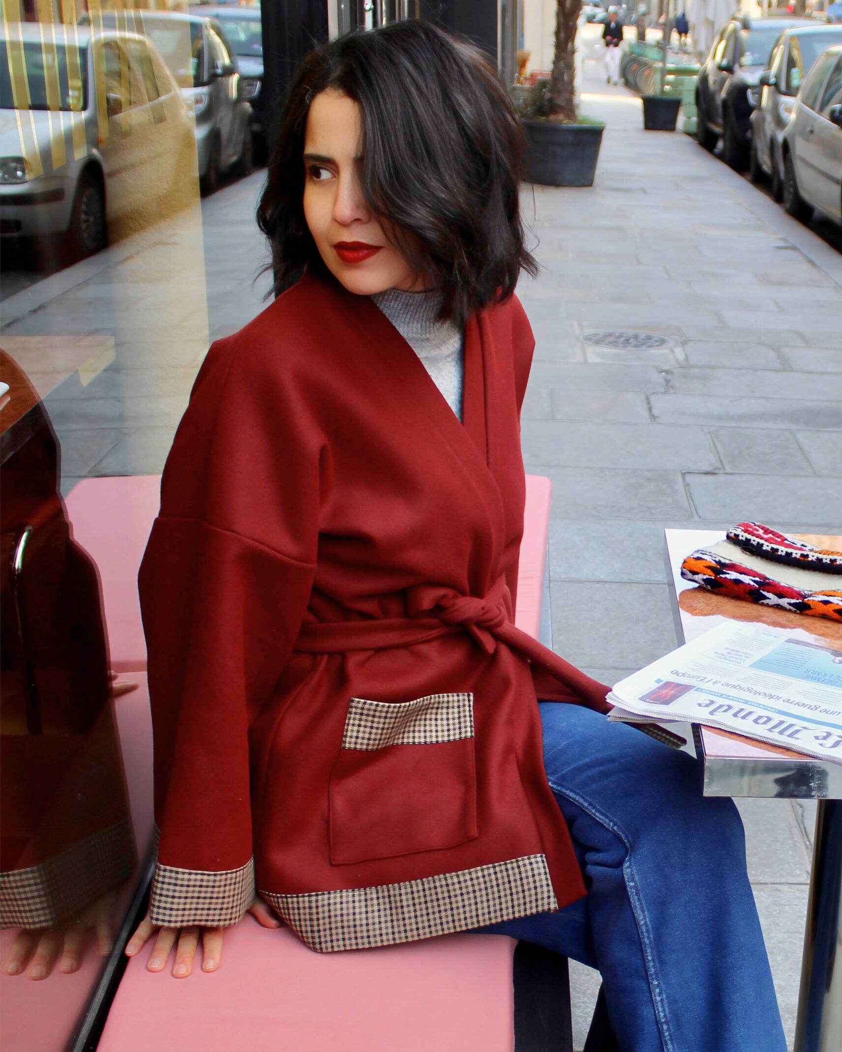 Jeune femme à la terrasse d'un café parisien, portant un kimono en laine bordeaux et finitions en laine pied de poule.