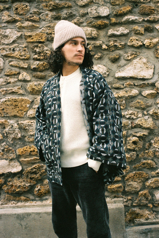 Homme portant le kimono VERDEAU Atelier Sarita, veste en jacquard géométrique bleu, gris et kaki, avec grandes poches plaquées. Kimono fabriqué à la main à Paris dans un jacquard issu d’une maison textile française historique. Photographie en extérieur devant un mur en pierre, mettant en valeur la texture dense du jacquard et la coupe ample et structurée du modèle.