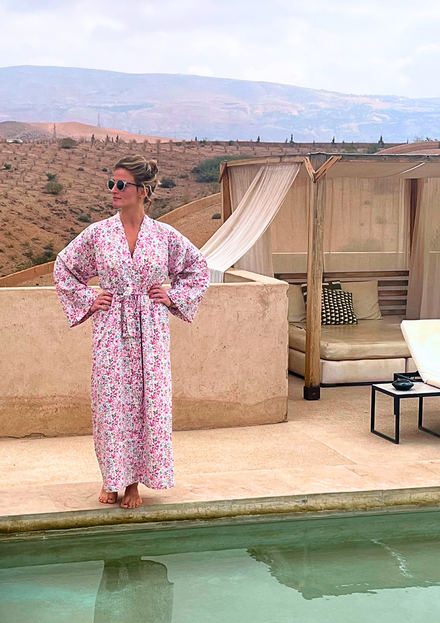 Femme portant un kimono long en coton fleuri rose Atelier Sarita, debout au bord d'une piscine dans un paysage désertique.