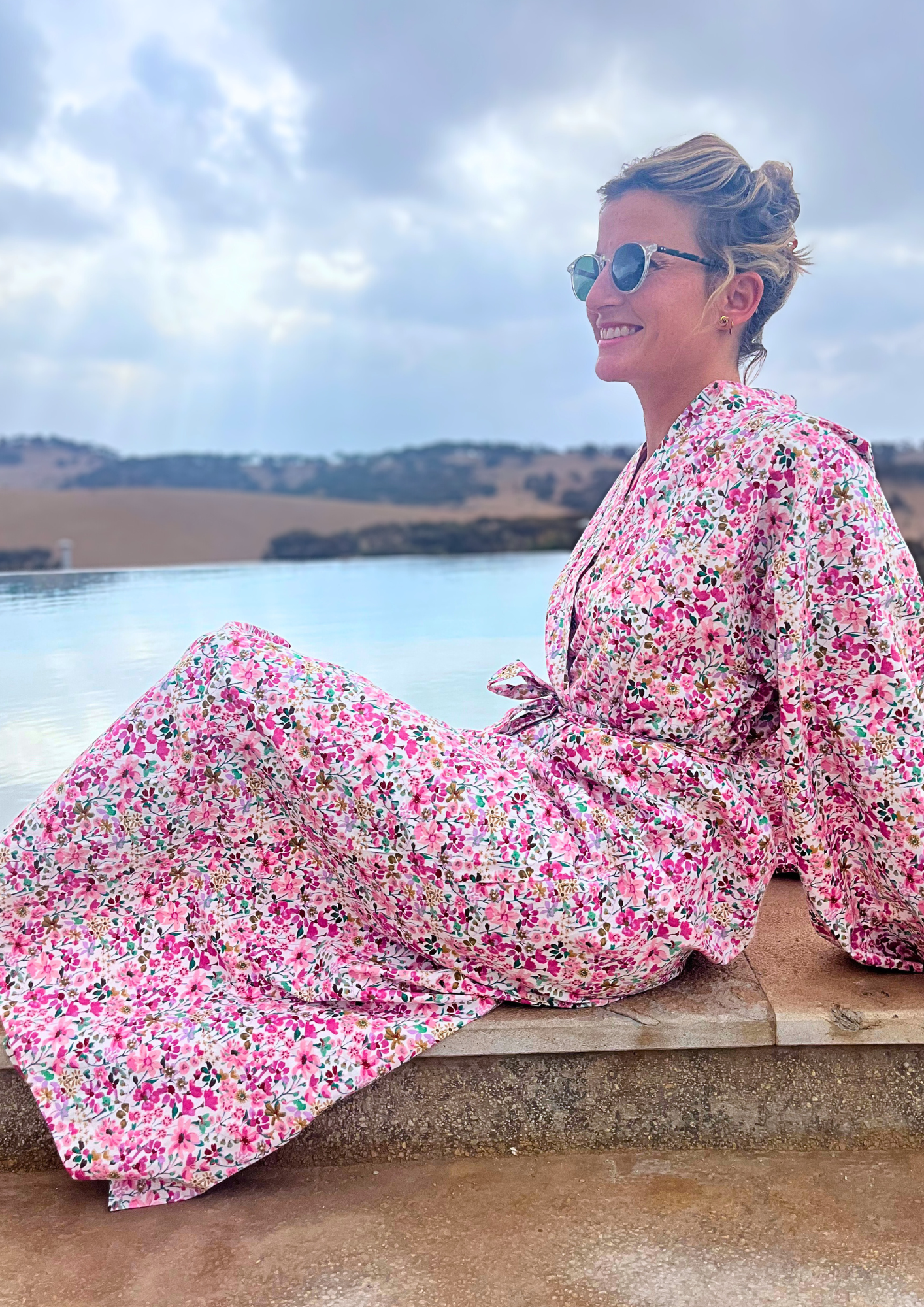 Femme portant un kimono long en coton fleuri rose Atelier Sarita, debout au bord d'une piscine dans un paysage désertique.