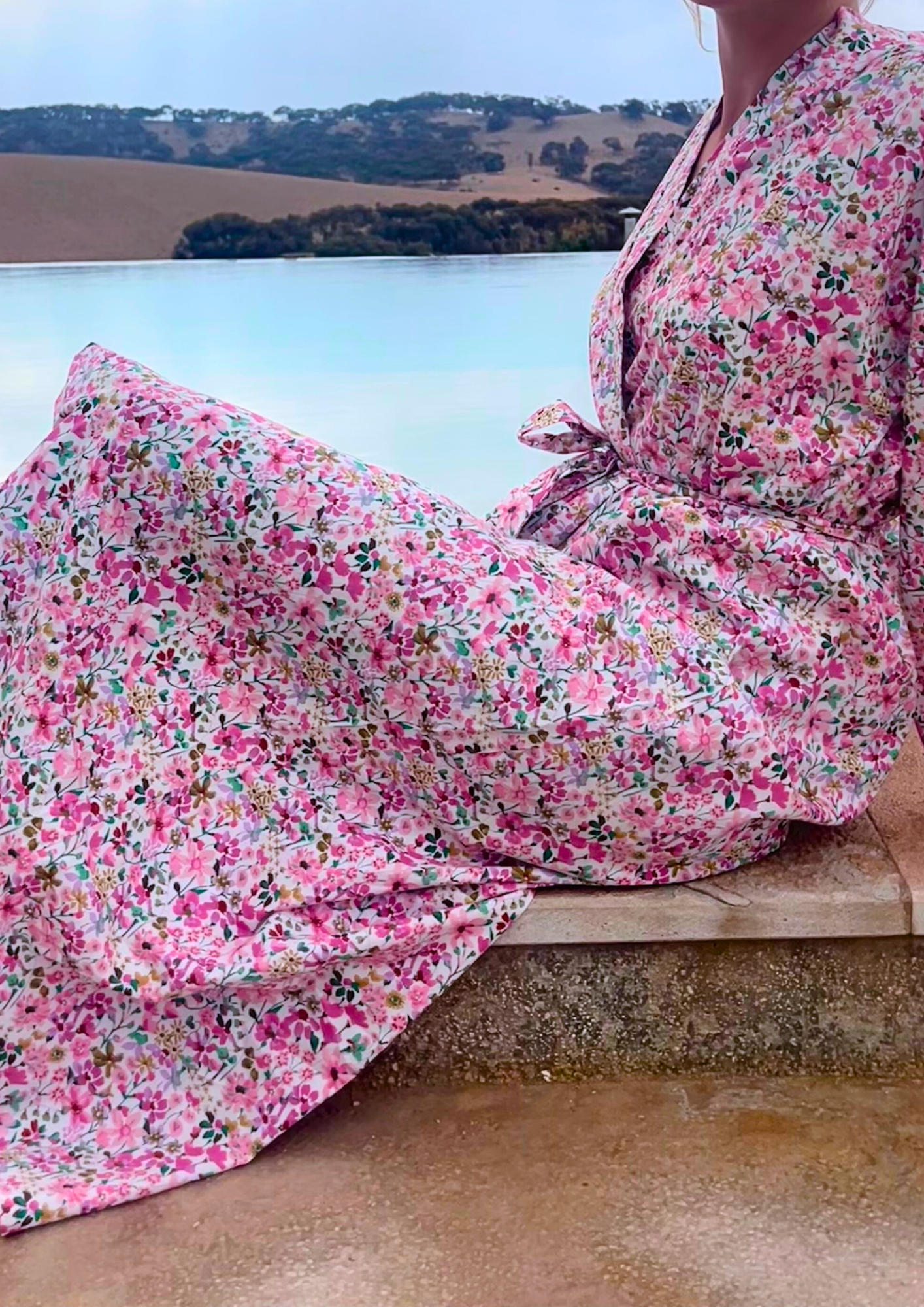 Femme portant un kimono long en coton fleuri rose Atelier Sarita, debout au bord d'une piscine dans un paysage désertique.