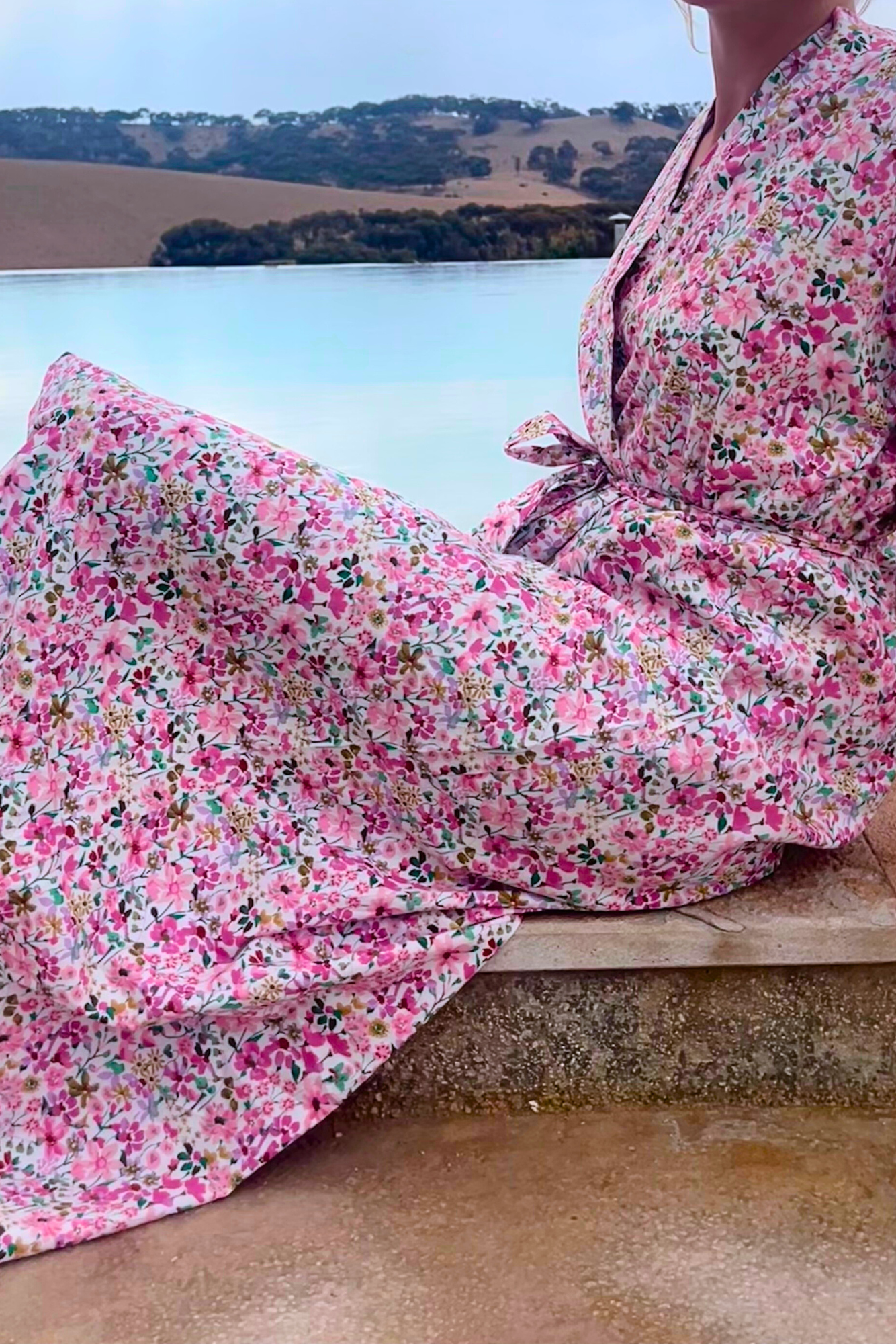 Femme portant un kimono long en coton fleuri rose Atelier Sarita, debout au bord d'une piscine dans un paysage désertique.