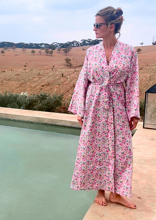 Femme portant un kimono long en coton fleuri rose Atelier Sarita, debout au bord d'une piscine dans un paysage désertique.