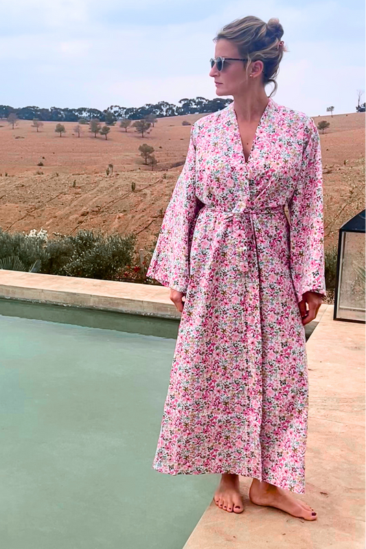 Femme portant un kimono long en coton fleuri rose Atelier Sarita, debout au bord d'une piscine dans un paysage désertique.