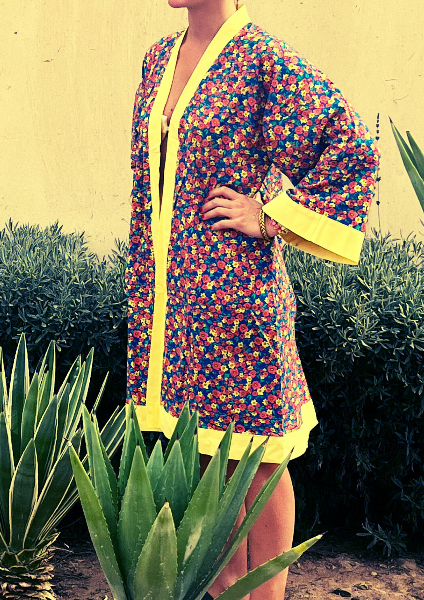 Kimono fleuri Olinda à finitions jaunes, coupe courte estivale, porté devant un mur sable et des plantes tropicales.