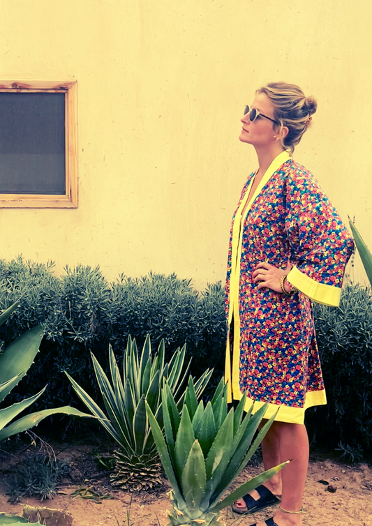 Kimono fleuri Olinda à finitions jaunes, coupe courte estivale, porté devant un mur sable et des plantes tropicales.