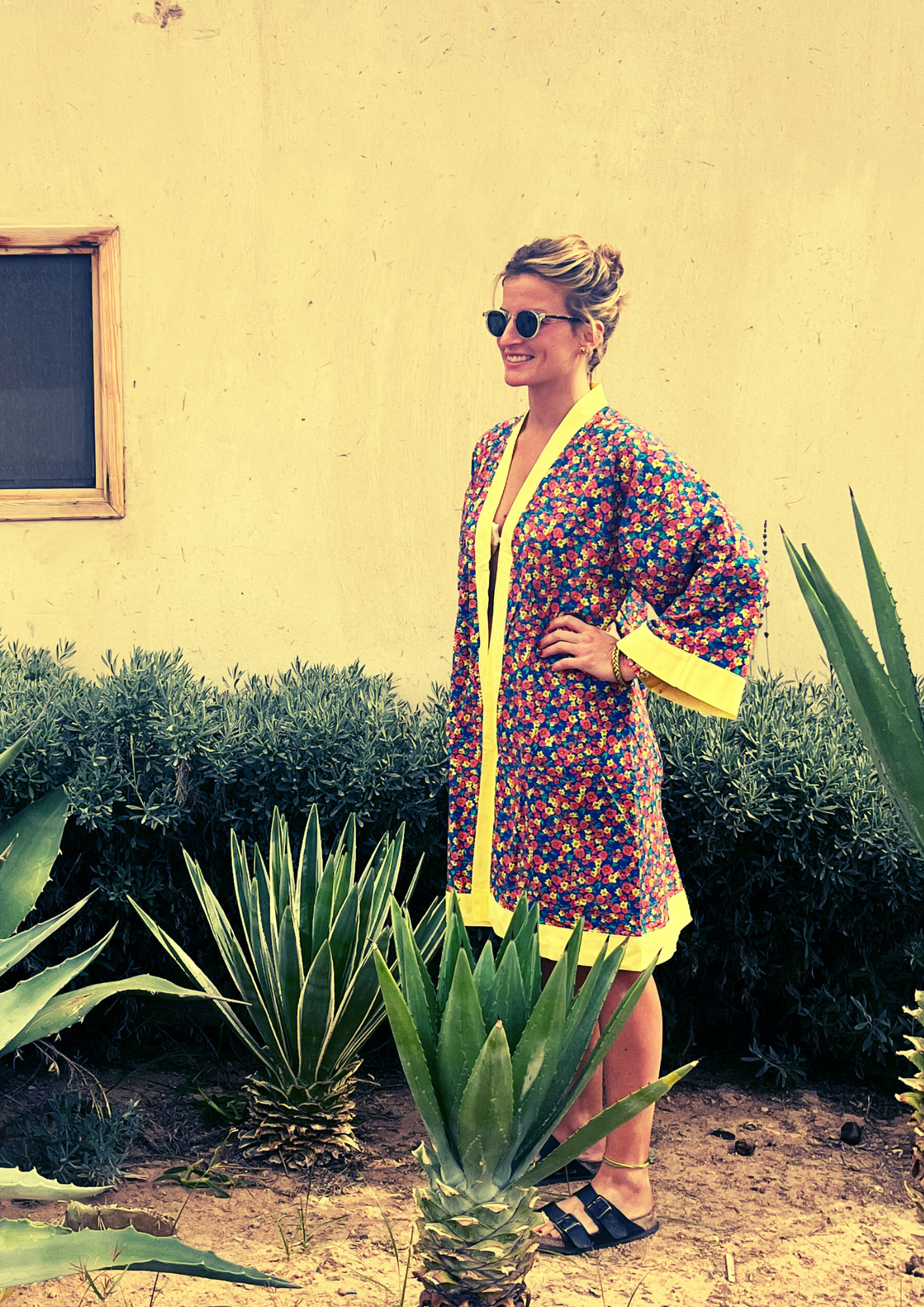 Kimono fleuri Olinda à finitions jaunes, coupe courte estivale, porté devant un mur sable et des plantes tropicales.