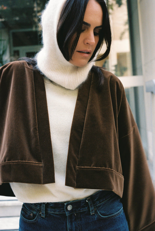Femme portant le kimono court BRADY en velours de coton marron café Atelier Sarita avec poches intégrées. Kimono fabriqué à la main à Paris dans un velours 100 % coton issu de deadstocks. Pièce chaude et texturée, au tombé souple, photographiée en extérieur avec un look hivernal composé d’un pull écru et d’un jean, mettant en valeur la douceur et la profondeur du velours marron.