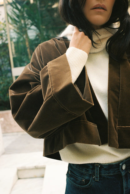 Femme portant le kimono court BRADY en velours de coton marron café Atelier Sarita avec poches intégrées. Kimono fabriqué à la main à Paris dans un velours 100 % coton issu de deadstocks. Pièce chaude et texturée, au tombé souple, photographiée en extérieur avec un look hivernal composé d’un pull écru et d’un jean, mettant en valeur la douceur et la profondeur du velours marron.