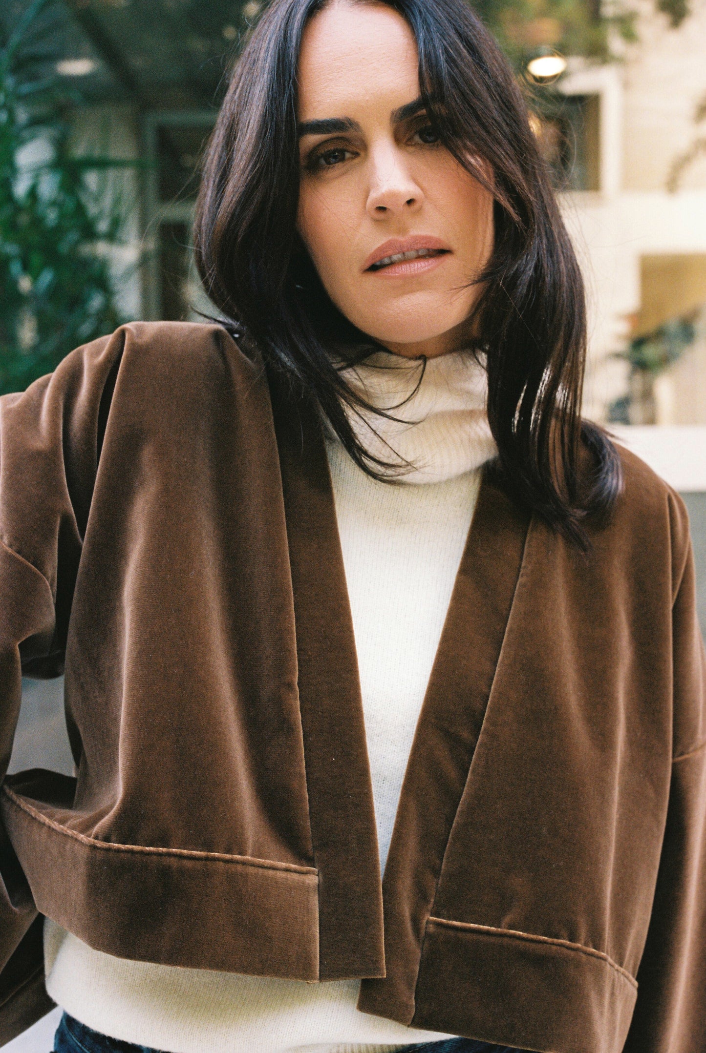 Femme portant le kimono court BRADY en velours de coton marron café Atelier Sarita avec poches intégrées. Kimono fabriqué à la main à Paris dans un velours 100 % coton issu de deadstocks. Pièce chaude et texturée, au tombé souple, photographiée en extérieur avec un look hivernal composé d’un pull écru et d’un jean, mettant en valeur la douceur et la profondeur du velours marron.