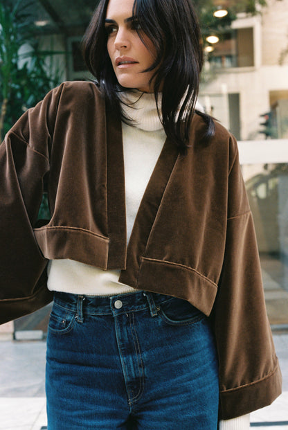 Femme portant le kimono court BRADY en velours de coton marron café Atelier Sarita avec poches intégrées. Kimono fabriqué à la main à Paris dans un velours 100 % coton issu de deadstocks. Pièce chaude et texturée, au tombé souple, photographiée en extérieur avec un look hivernal composé d’un pull écru et d’un jean, mettant en valeur la douceur et la profondeur du velours marron.