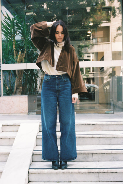 Femme portant le kimono court BRADY en velours de coton marron café Atelier Sarita avec poches intégrées. Kimono fabriqué à la main à Paris dans un velours 100 % coton issu de deadstocks. Pièce chaude et texturée, au tombé souple, photographiée en extérieur avec un look hivernal composé d’un pull écru et d’un jean, mettant en valeur la douceur et la profondeur du velours marron.