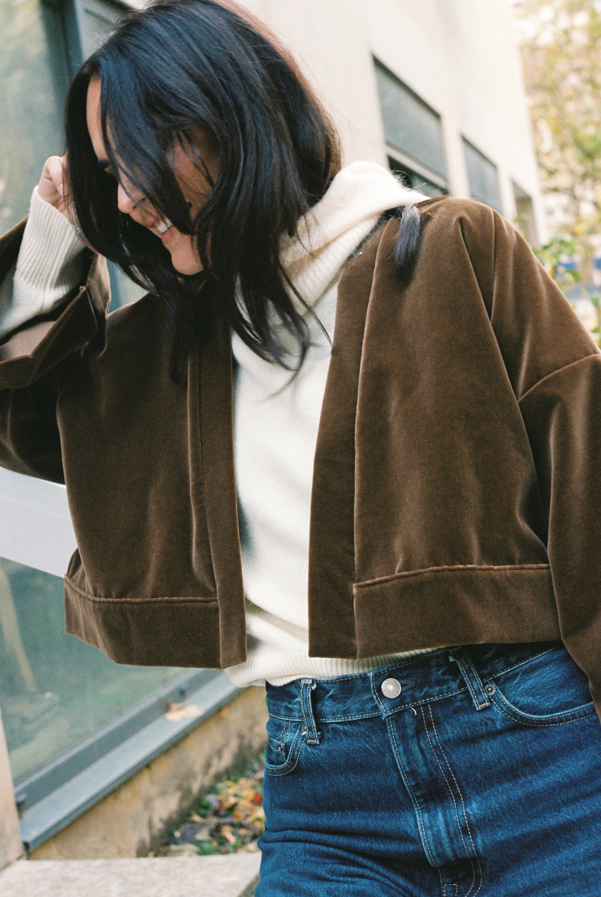 Femme portant le kimono court BRADY en velours de coton marron café Atelier Sarita avec poches intégrées. Kimono fabriqué à la main à Paris dans un velours 100 % coton issu de deadstocks. Pièce chaude et texturée, au tombé souple, photographiée en extérieur avec un look hivernal composé d’un pull écru et d’un jean, mettant en valeur la douceur et la profondeur du velours marron.