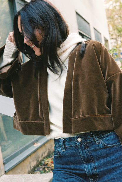 Femme portant le kimono court BRADY en velours de coton marron café Atelier Sarita avec poches intégrées. Kimono fabriqué à la main à Paris dans un velours 100 % coton issu de deadstocks. Pièce chaude et texturée, au tombé souple, photographiée en extérieur avec un look hivernal composé d’un pull écru et d’un jean, mettant en valeur la douceur et la profondeur du velours marron.