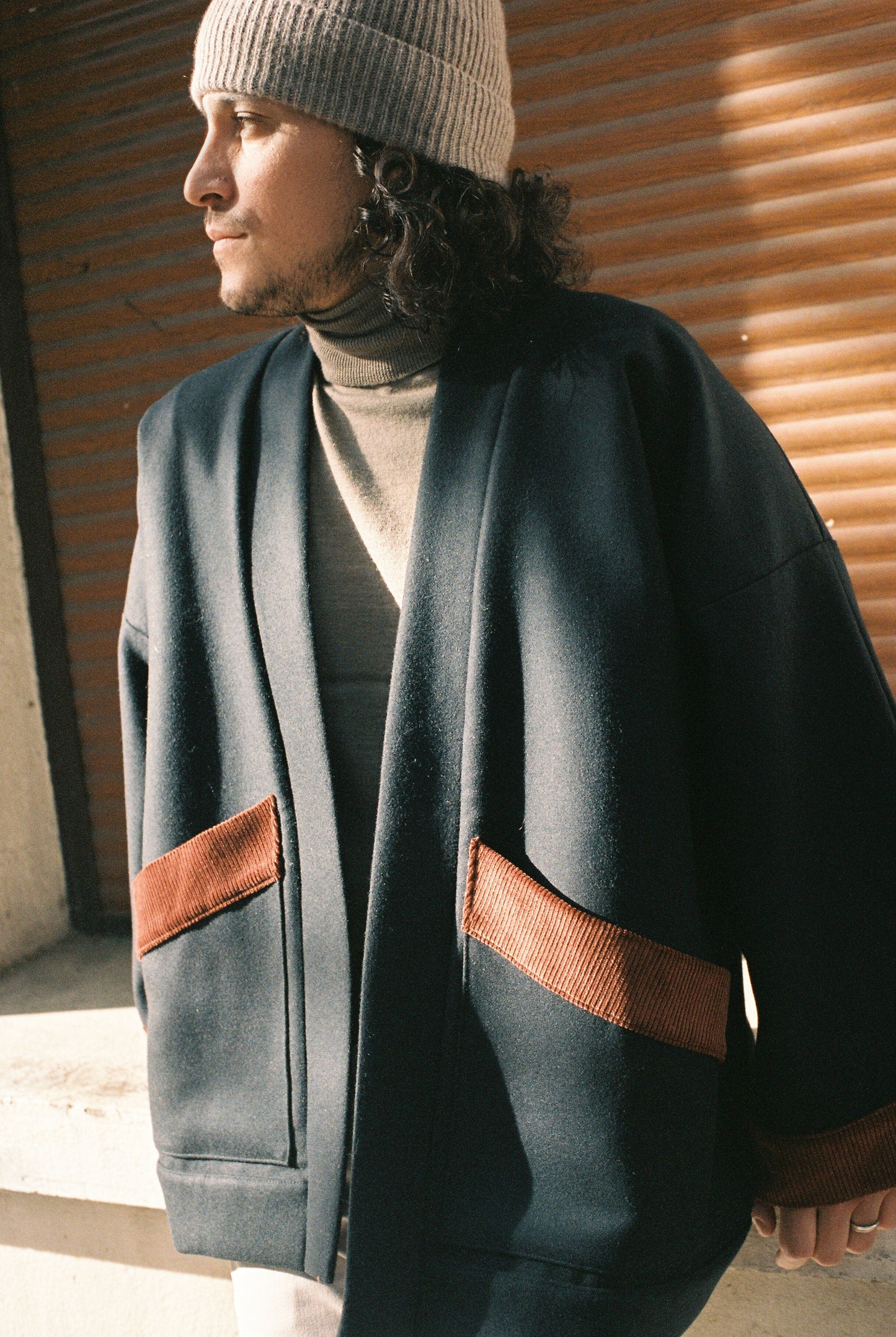 Homme portant le kimono DODAT Atelier Sarita, un kimono veste en laine caviar noire avec finitions en velours coton couleur oud, photographié en extérieur au soleil. Modèle unisexe, confectionné à la main à Paris, au style urbain et architectural.