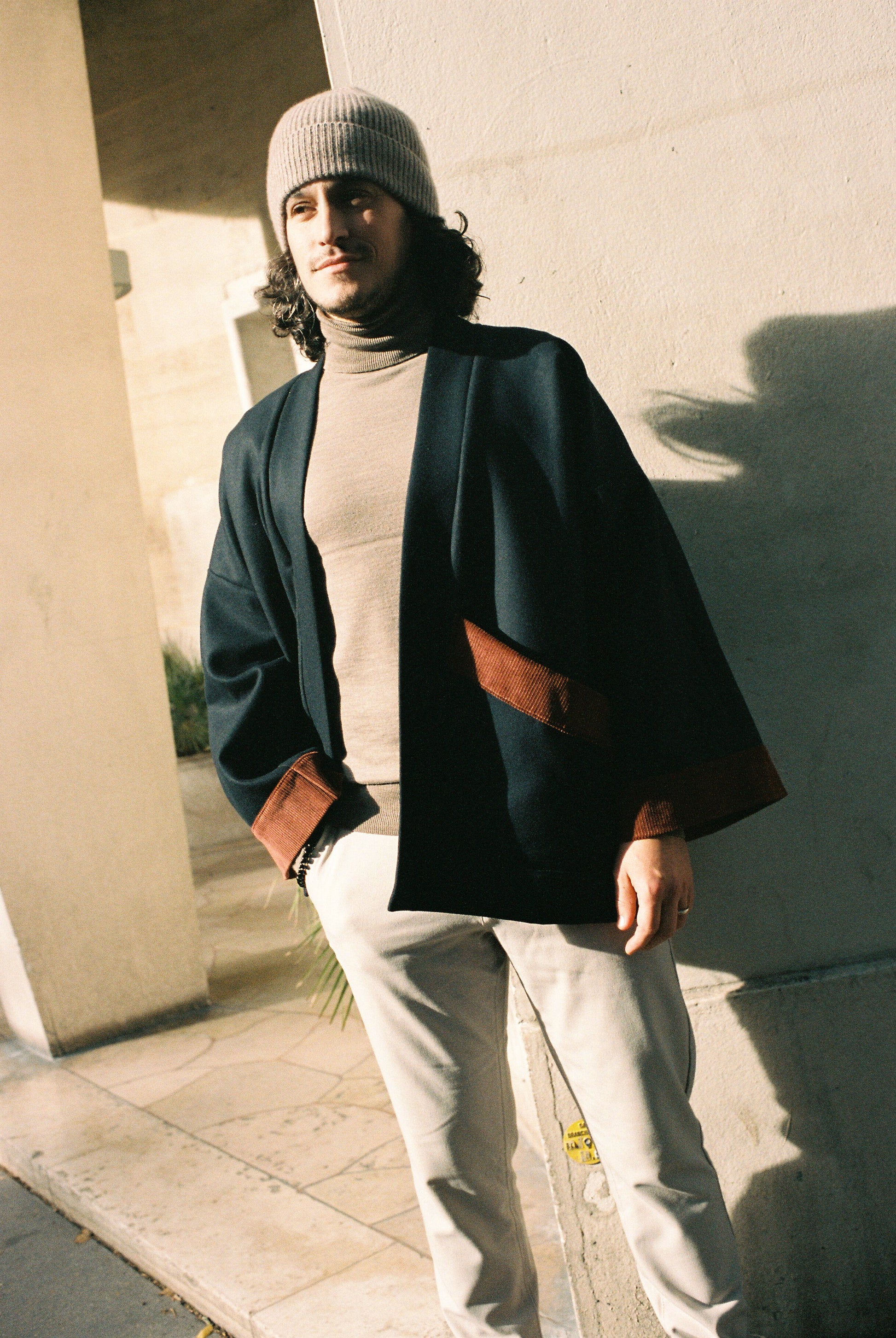 Homme portant le kimono DODAT Atelier Sarita, un kimono veste en laine caviar noire avec finitions en velours coton couleur oud, photographié en extérieur au soleil. Modèle unisexe, confectionné à la main à Paris, au style urbain et architectural.