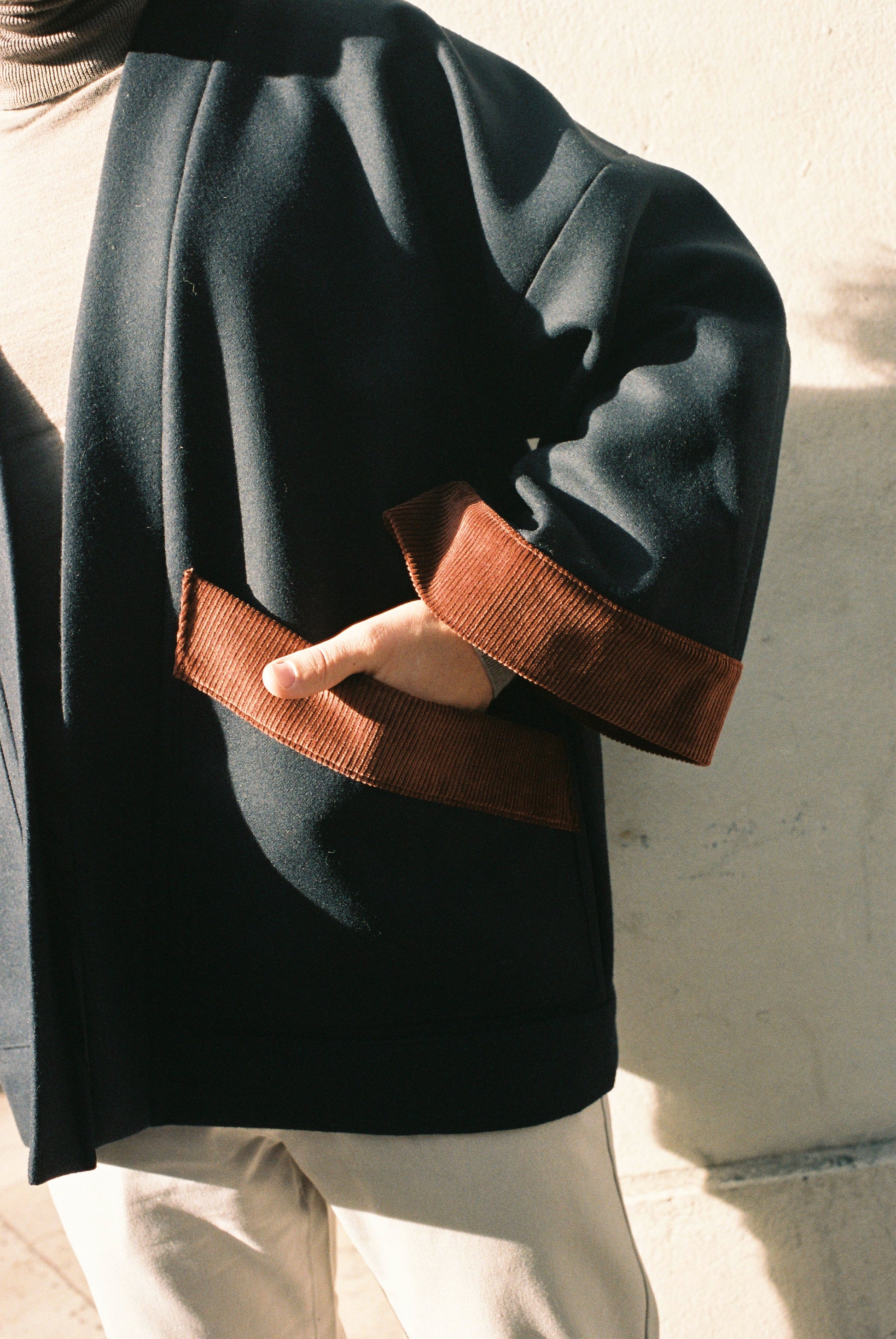 Personne portant le kimono DODAT Atelier Sarita, un kimono veste en laine caviar noire avec finitions en velours coton couleur oud, photographié en extérieur au soleil. Modèle unisexe, confectionné à la main à Paris, au style urbain et architectural.