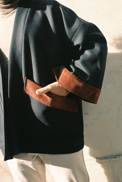 Personne portant le kimono DODAT Atelier Sarita, un kimono veste en laine caviar noire avec finitions en velours coton couleur oud, photographié en extérieur au soleil. Modèle unisexe, confectionné à la main à Paris, au style urbain et architectural.