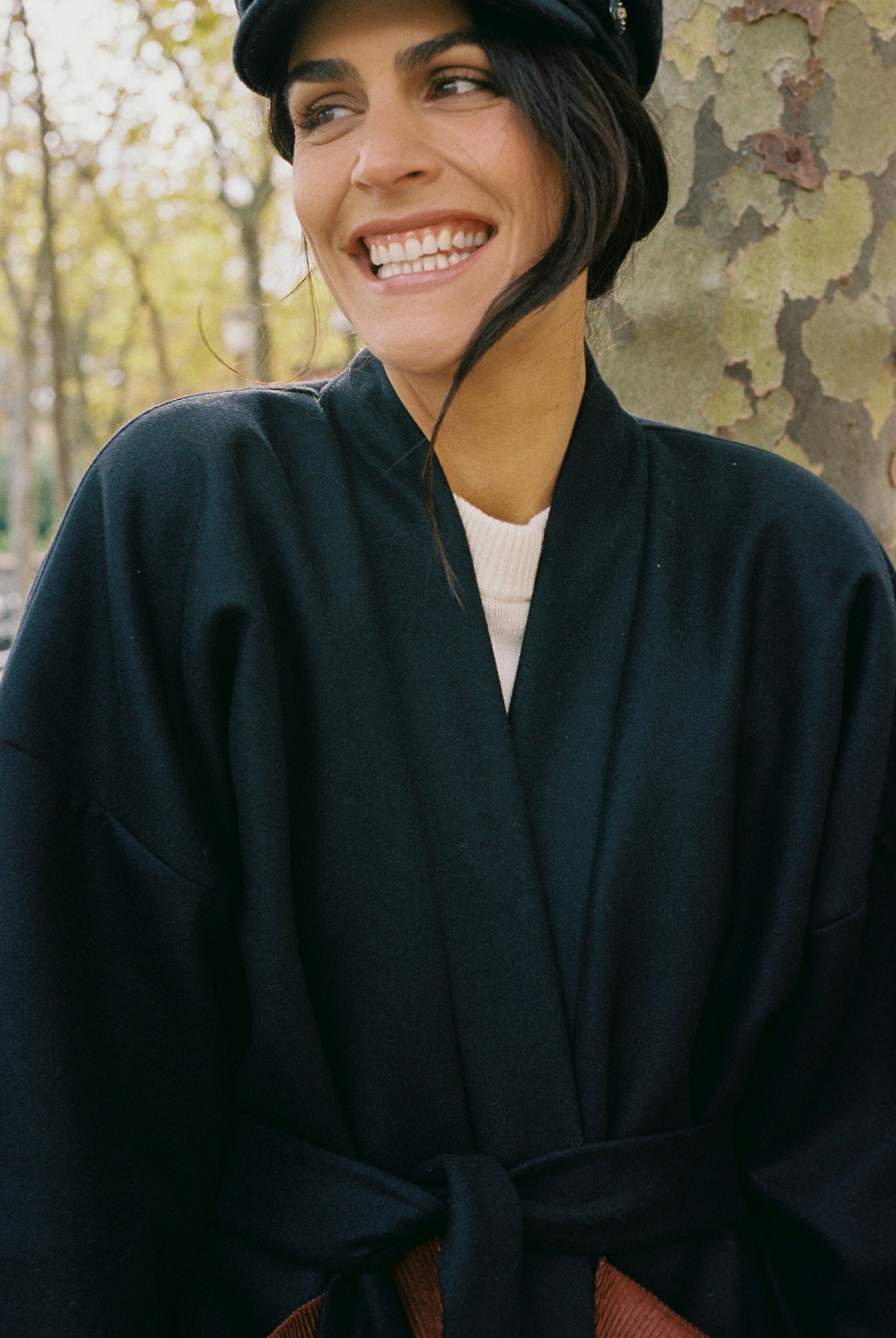 Femme portant le kimono DODAT Atelier Sarita, un kimono veste en laine caviar noire avec finitions en velours coton couleur oud, photographié en extérieur au soleil. Modèle unisexe, confectionné à la main à Paris, au style urbain et architectural.