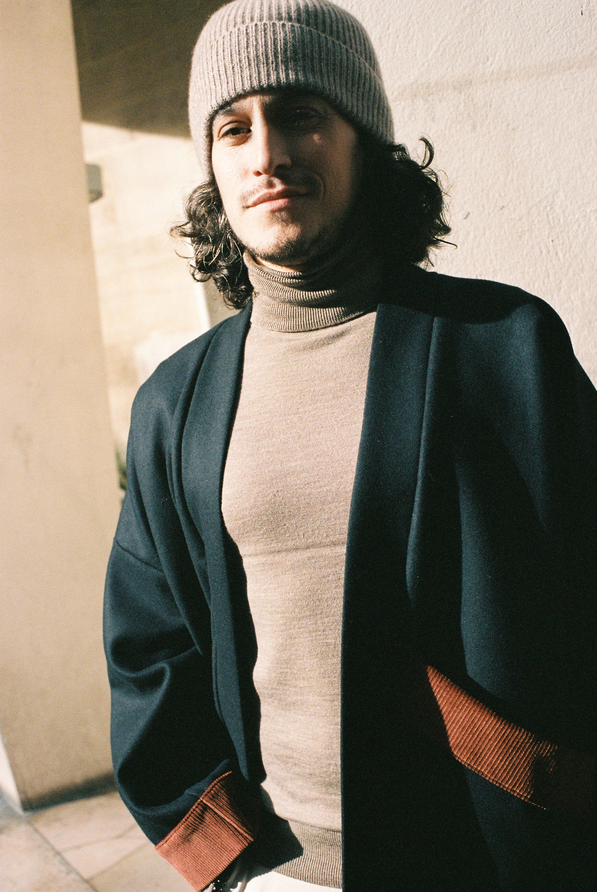 Homme portant le kimono DODAT Atelier Sarita, un kimono veste en laine caviar noire avec finitions en velours coton couleur oud, photographié en extérieur au soleil. Modèle unisexe, confectionné à la main à Paris, au style urbain et architectural.